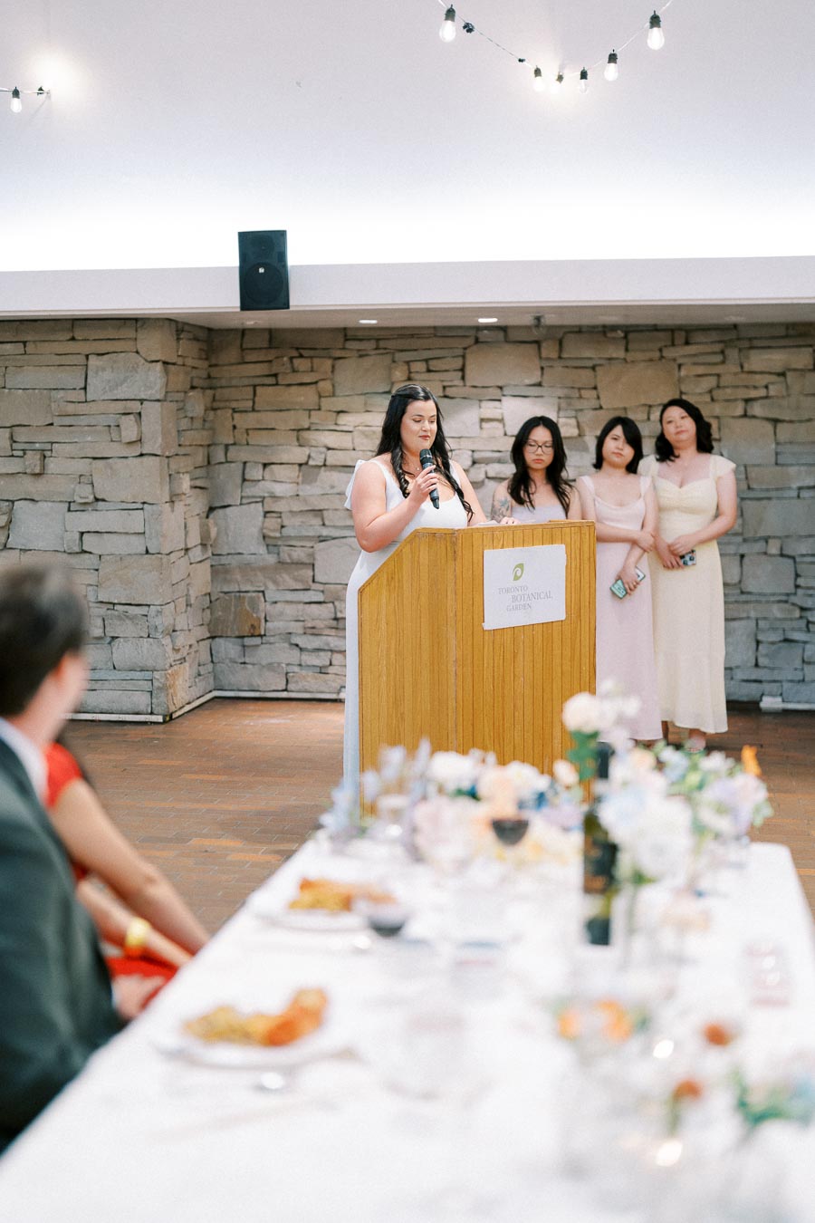 A woman delivers a speech at a podium during a formal event at the Toronto Botanical Garden, with three women standing behind her. The setting features a stone wall and a decorated table in the foreground.