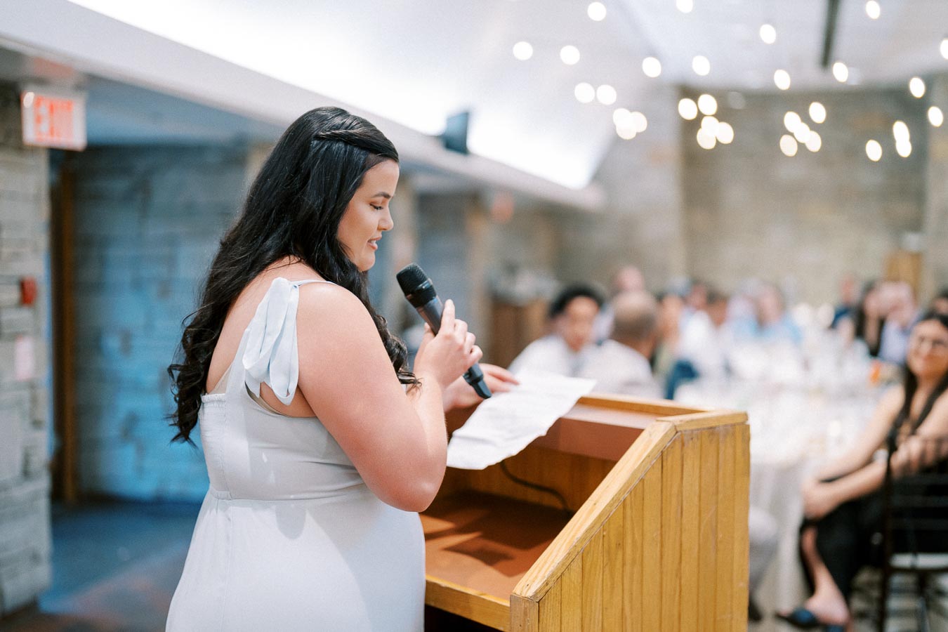 A woman in a light blue dress delivers a speech at a wooden podium during a formal event in a warmly lit venue with an audience in the background.