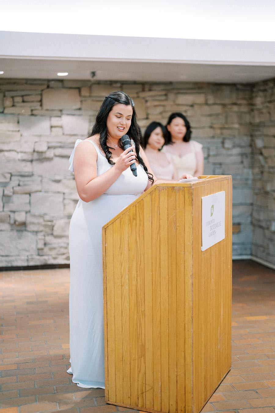 A woman delivers a speech at the Toronto Botanical Garden, standing at a wooden podium with two women in the background.