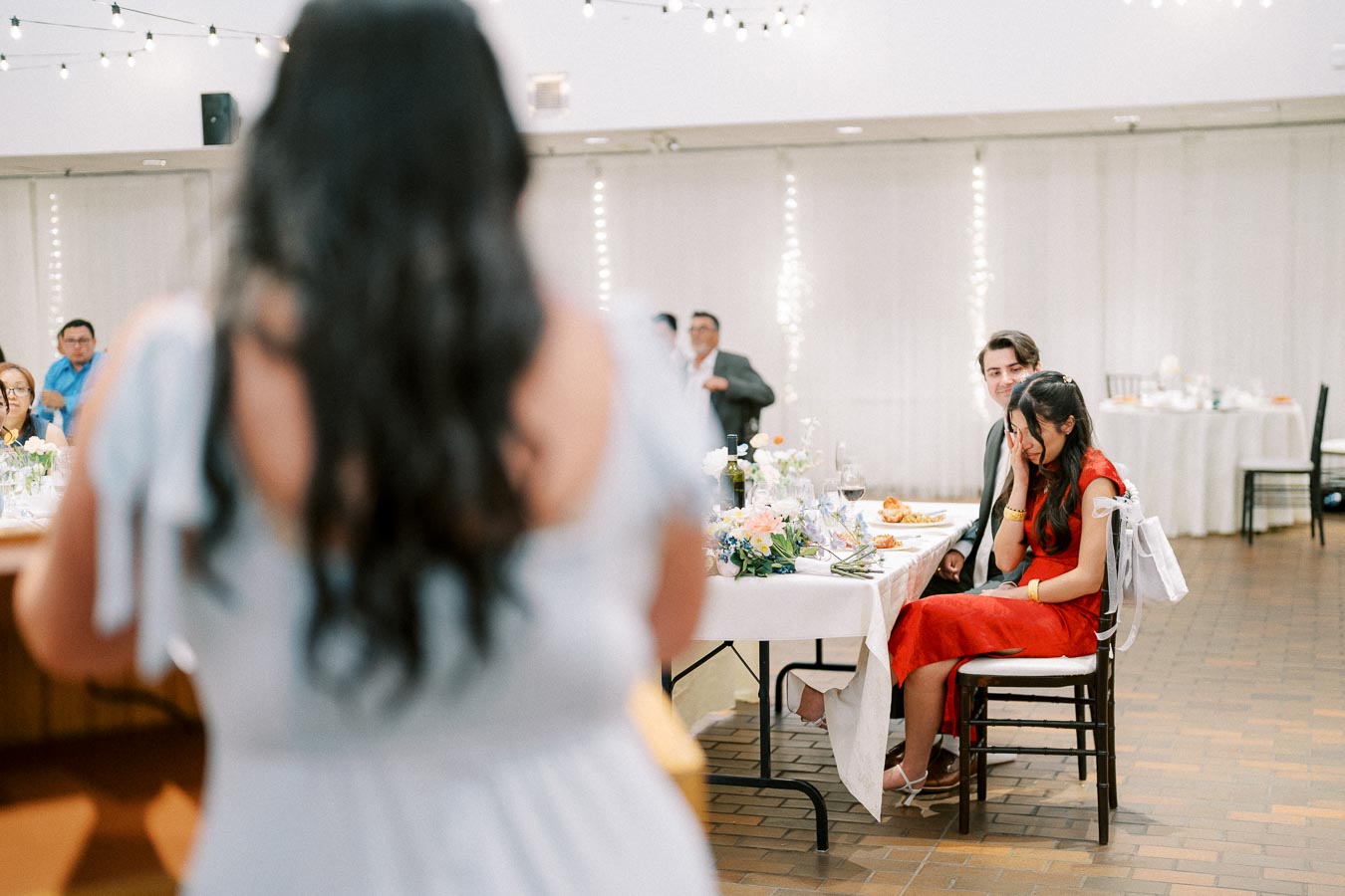 A joyful moment captured at a wedding reception, featuring a woman in a red dress and a man seated at a decorated table, listening to a speech.