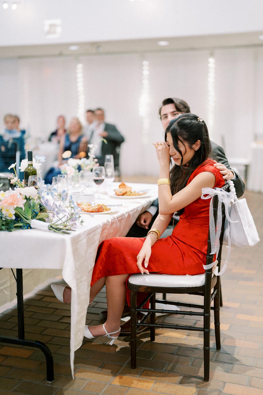 A woman in a red dress wipes her eyes at a formal event, comforted by a man beside her. The table is elegantly set with flowers and wine.