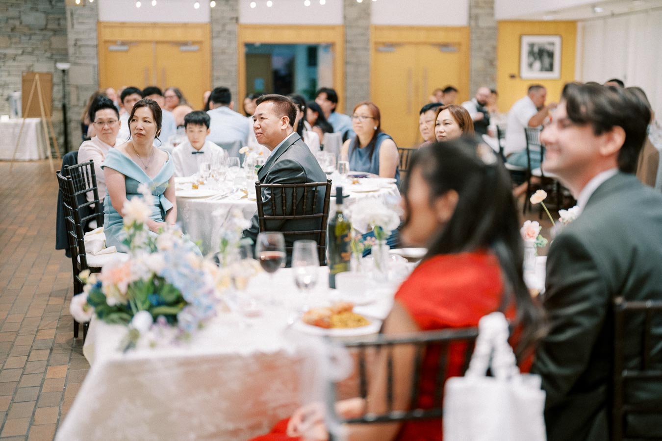 Guests seated at a formal event, attentively listening, surrounded by elegantly decorated tables.