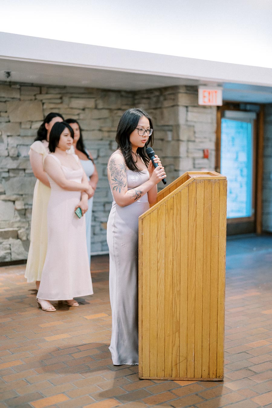 A woman delivers a speech at a podium during an indoor event, with three women standing behind her.