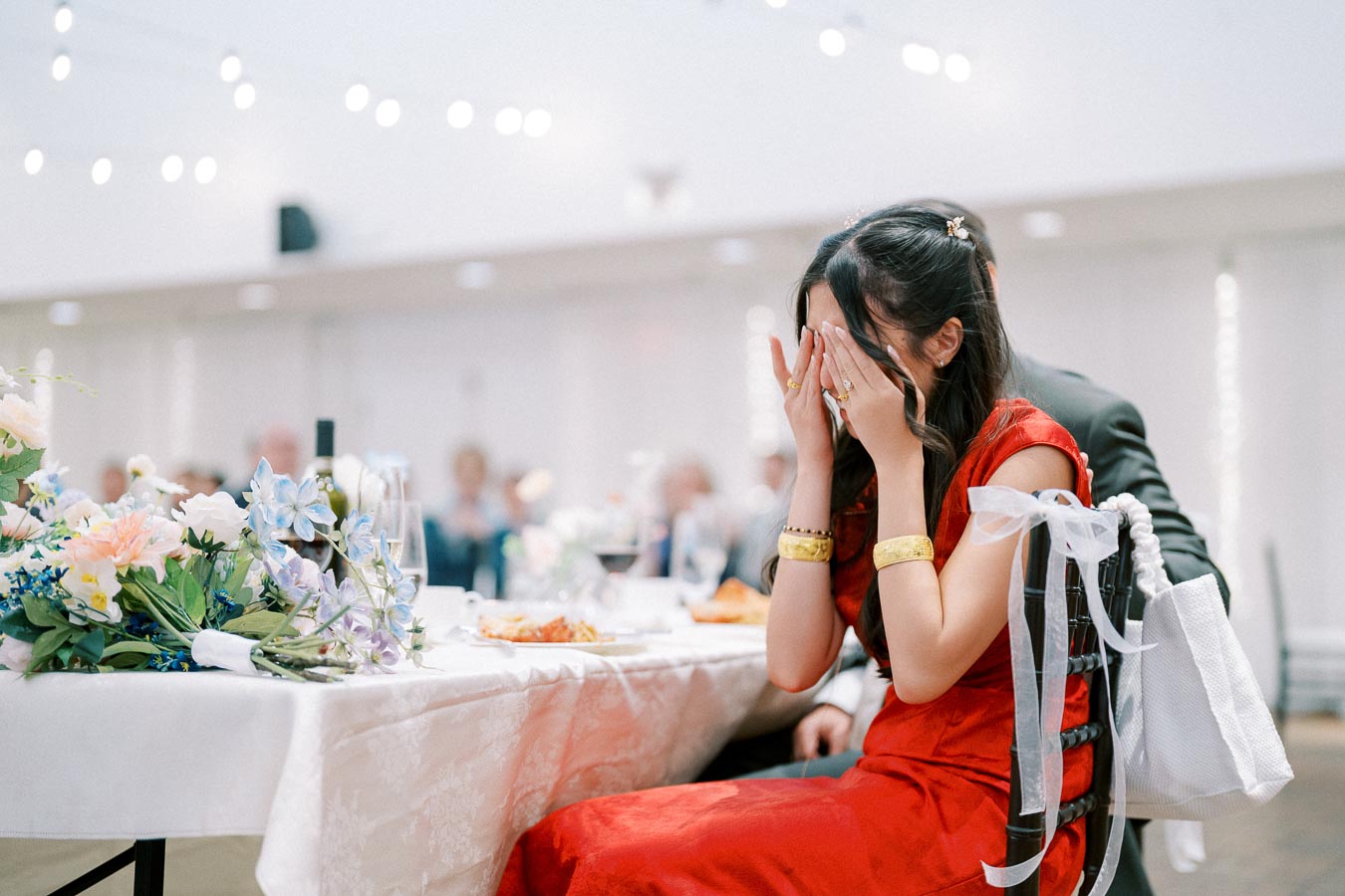 A woman in a red dress covers her face with her hands at a wedding reception, surrounded by floral decorations and soft lighting.