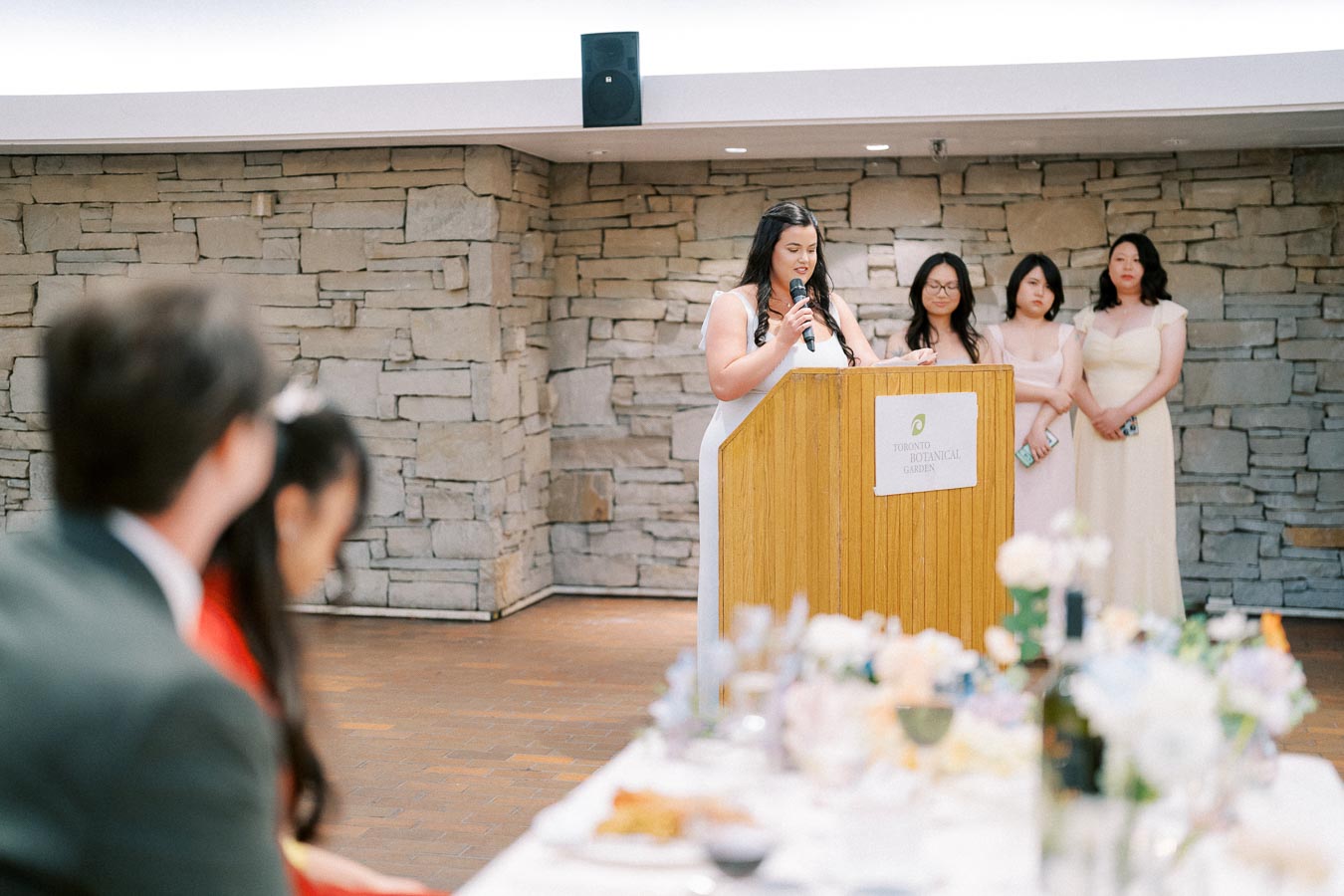 A woman in a light dress gives a speech at a podium marked Toronto Botanical Garden, accompanied by three women in similar attire, with a table set for a formal event in the foreground.