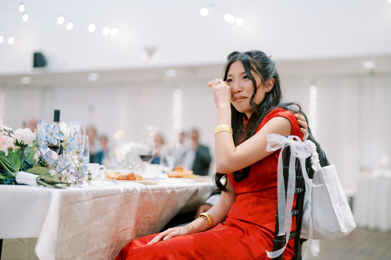 A woman in a red dress wipes her eye during a formal event, seated at a table adorned with flowers and wine glasses.