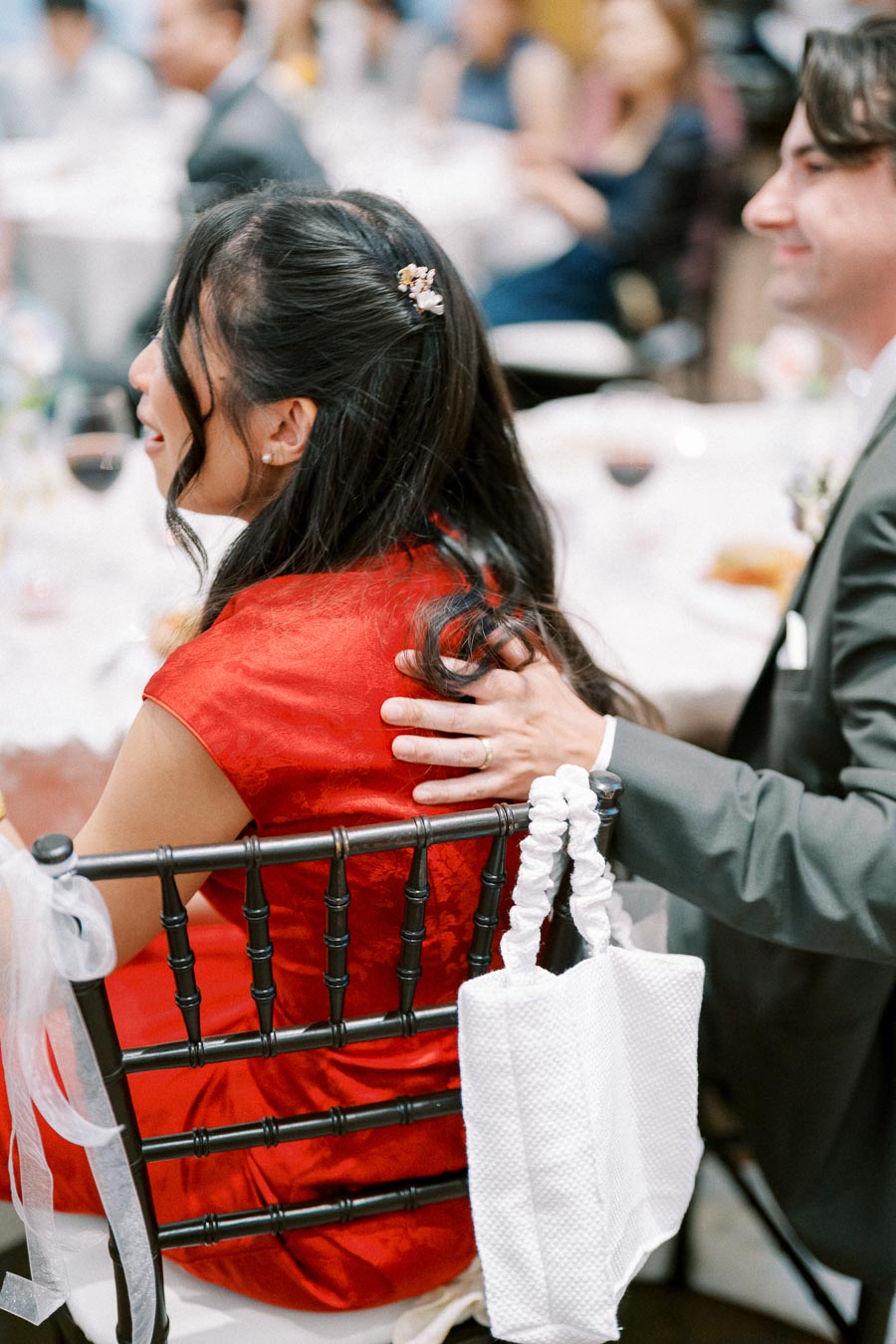 Wedding reception scene with a woman in a red dress sitting on a chair, adorned with a stylish hairpin, and a man in a suit with his hand gently resting on her shoulder. White cloth bag hanging from the chair.