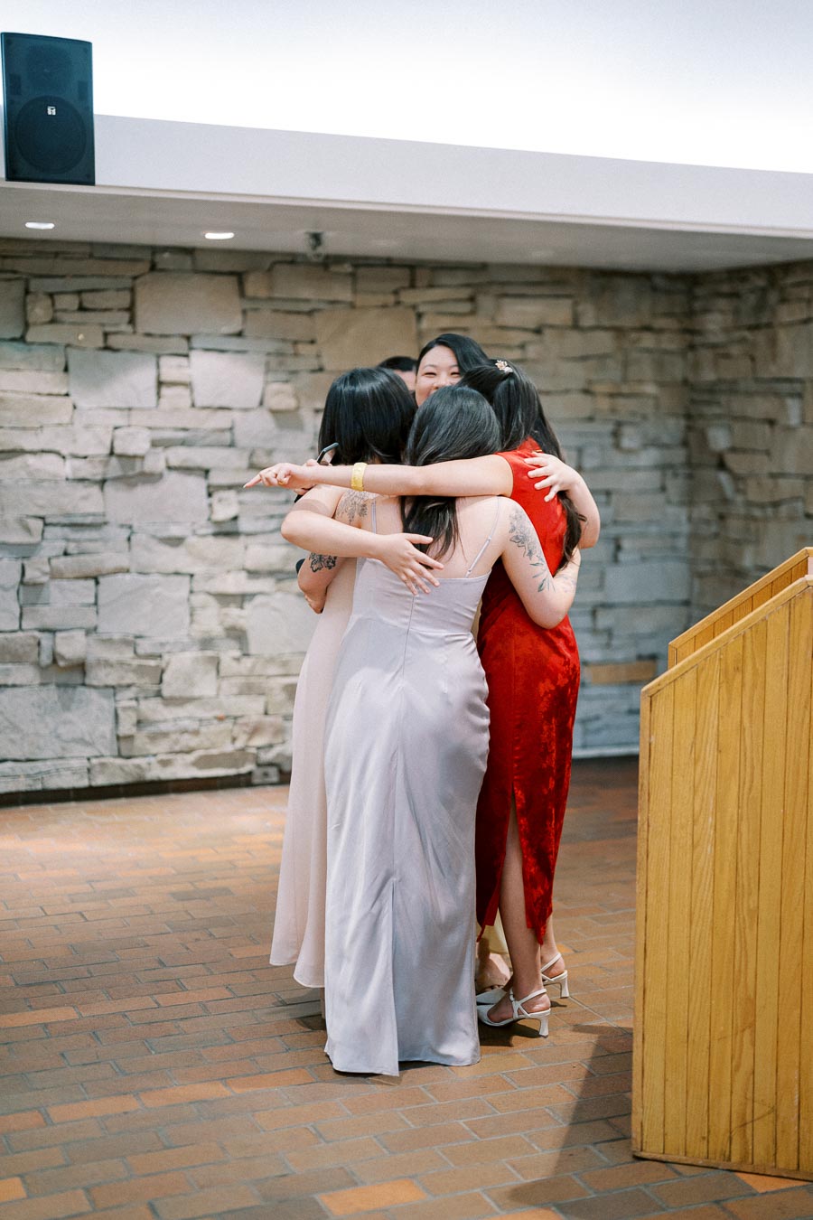 A group of women in elegant dresses hugging at an indoor event with stone wall background, celebrating friendship and connection.