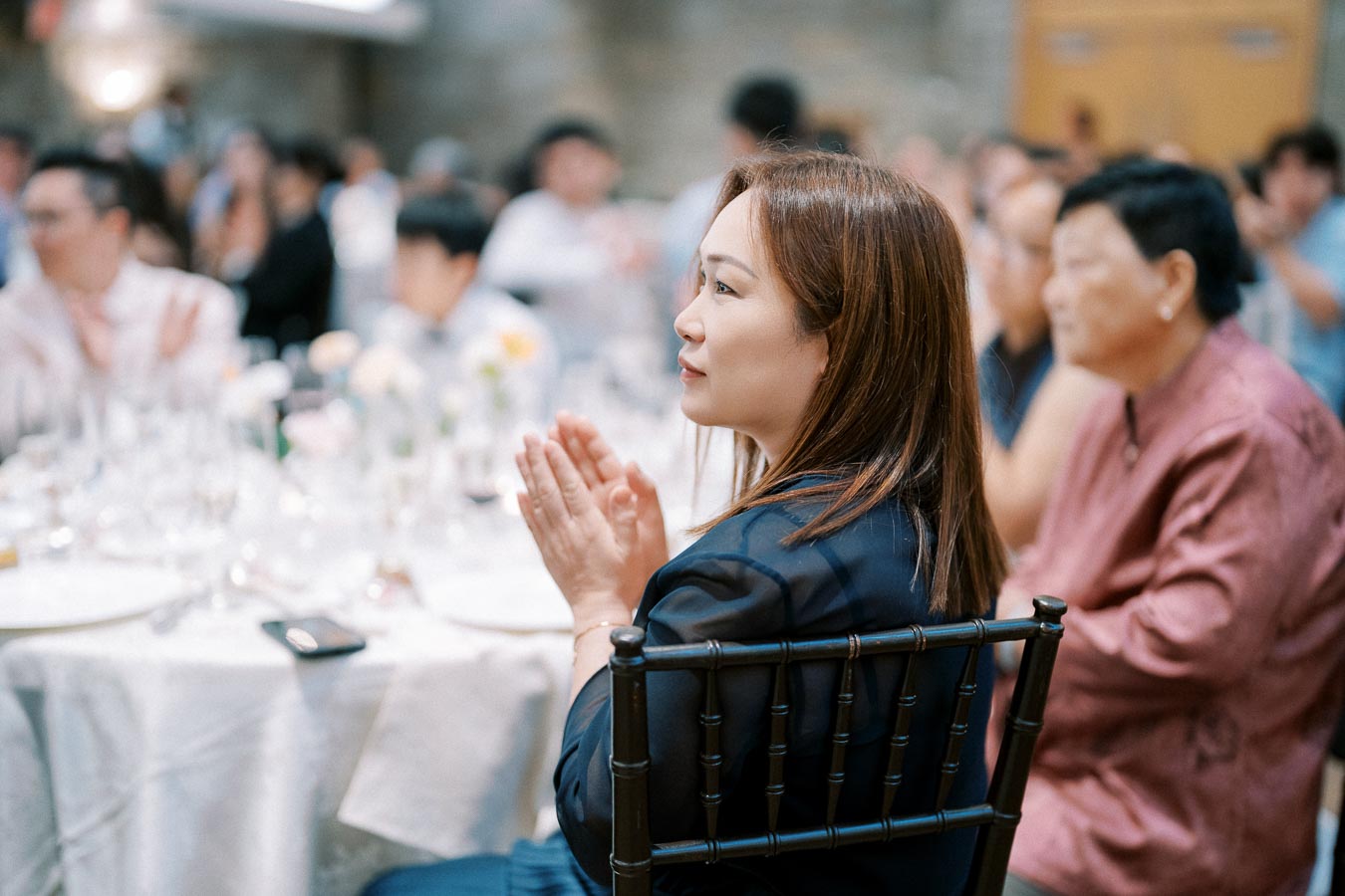A woman clapping at an indoor event, seated at a round table with a white tablecloth surrounded by a gathering of people, creating a lively and engaging atmosphere.