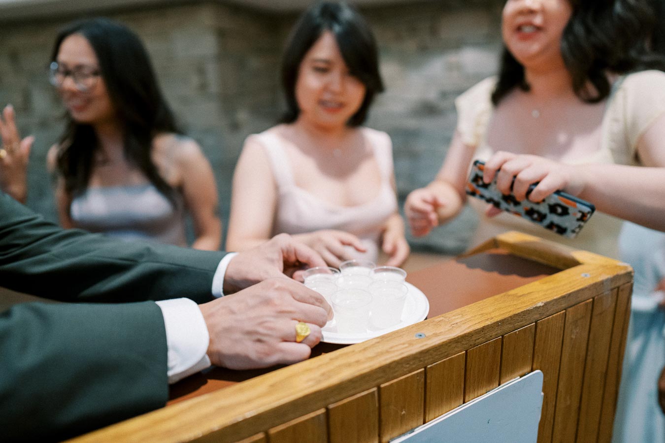 Group of people gathered around wooden bar counter, with hands reaching for small glasses on a tray at a social event.