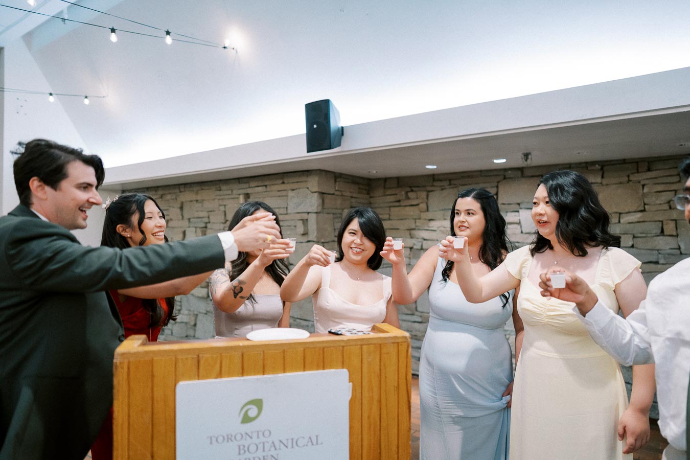 A group of people in formal attire cheerfully toasting with drinks at an indoor event at the Toronto Botanical Garden, surrounded by stone walls and decorative lighting.