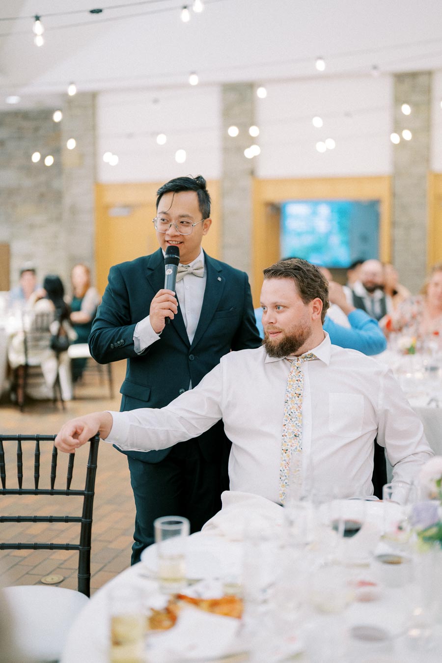 A man in a suit gives a speech at a formal event, standing next to a seated guest.