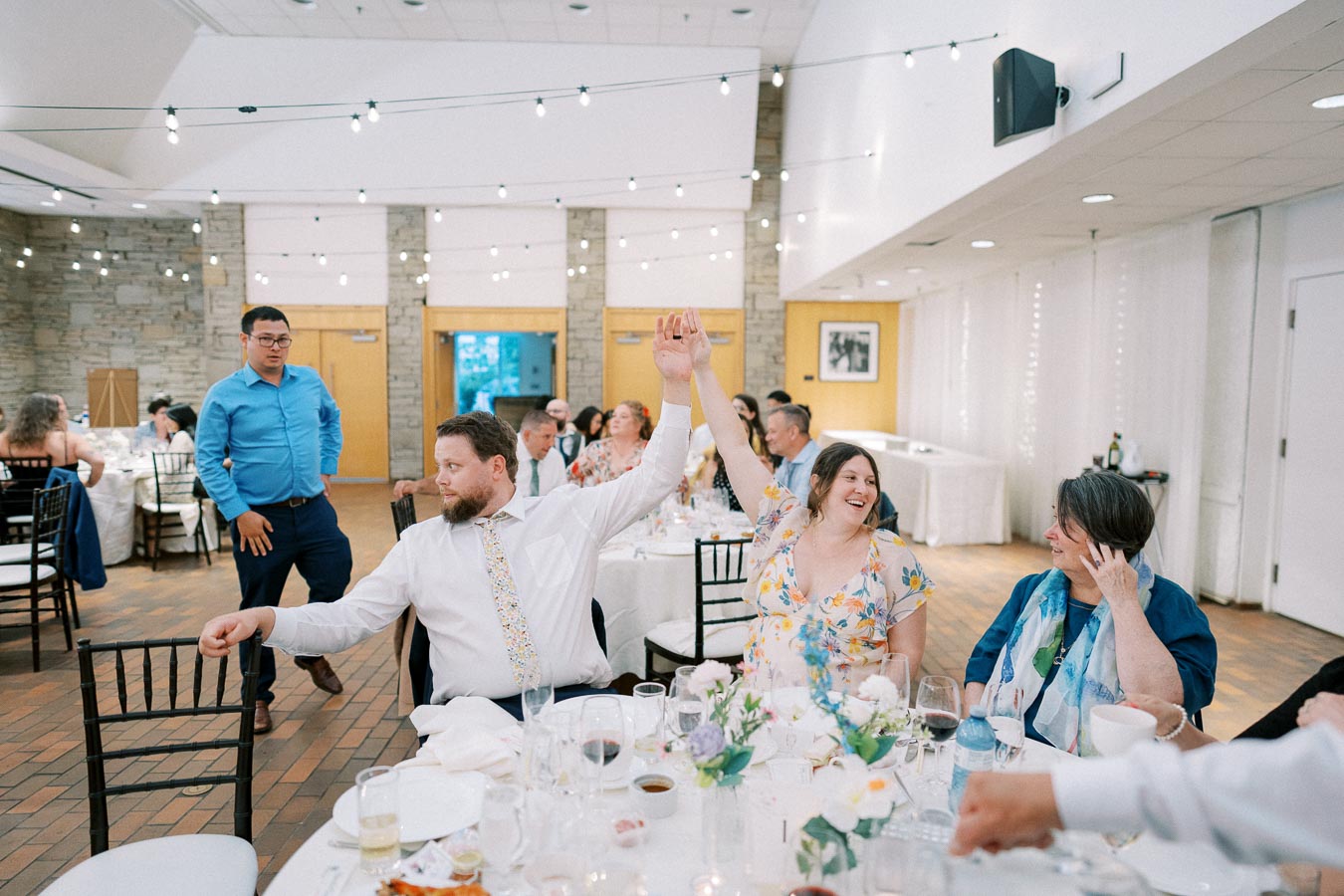 Guests enjoy a lively moment at a celebration, seated around a decorated table in a room adorned with string lights.