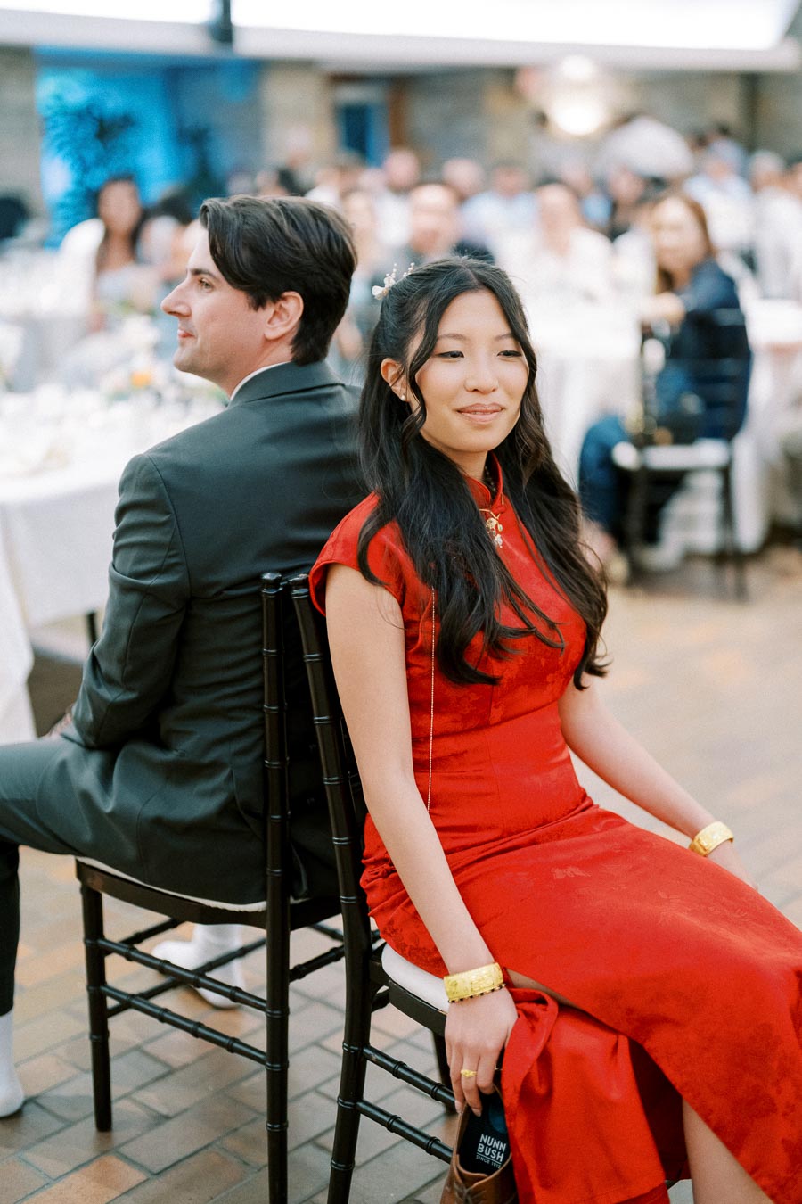 A couple participates in a wedding game, sitting back-to-back on chairs. The woman is dressed in a traditional red dress, while the man wears a suit. The setting appears to be a formal event with guests seated at tables in the background.