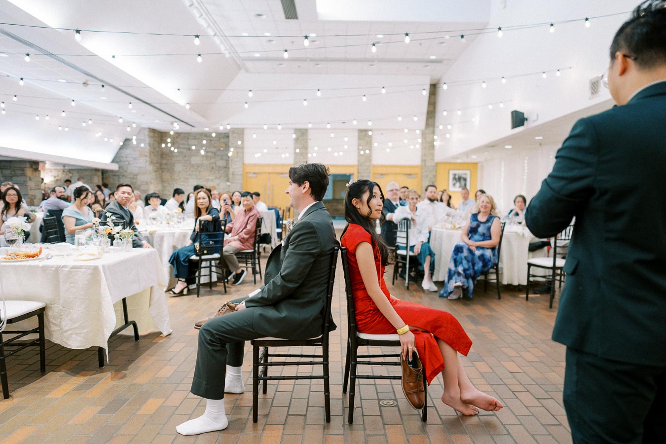 A couple participates in a fun wedding game, sitting back-to-back while holding shoes, as guests watch and enjoy the celebration.