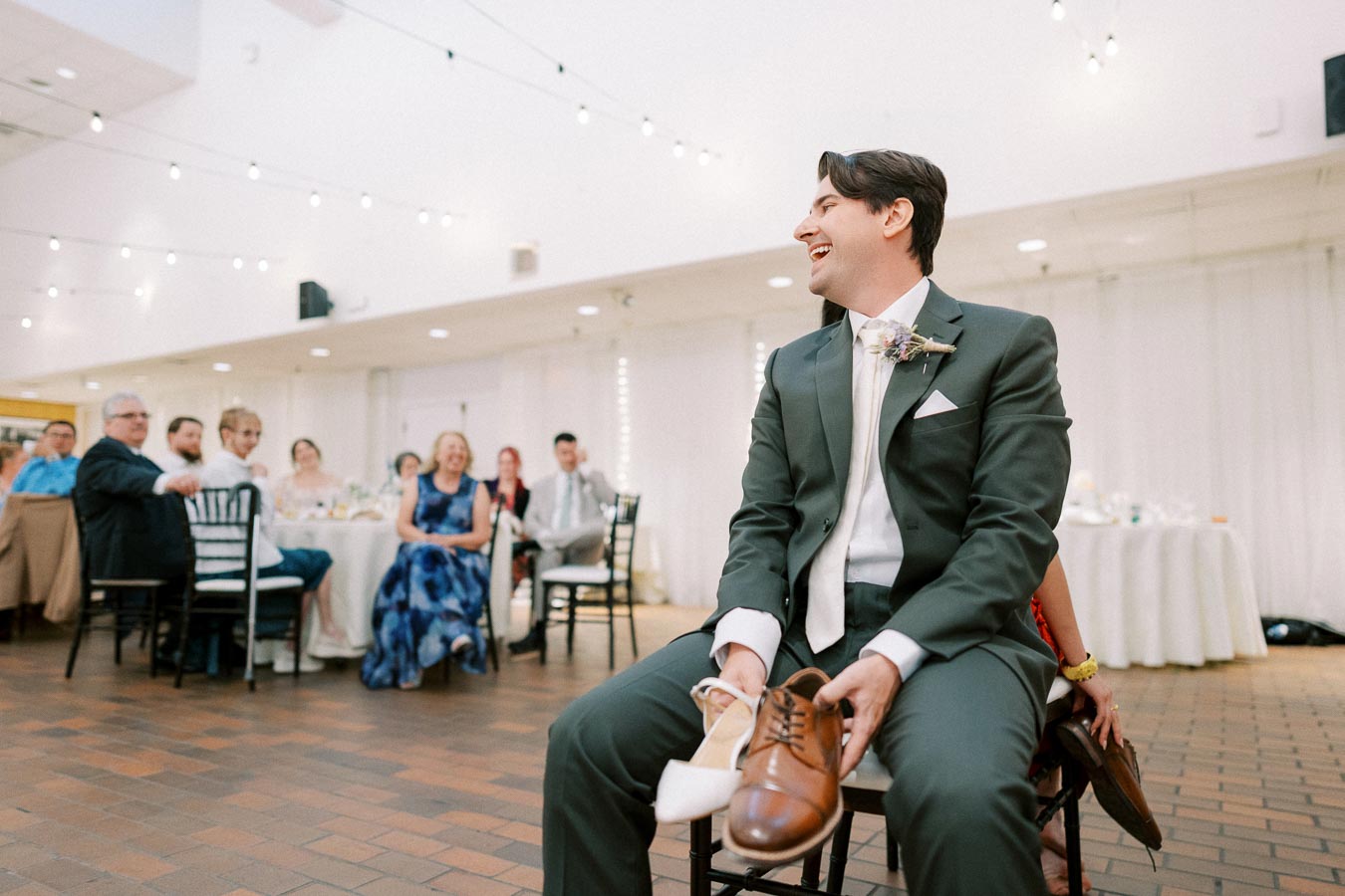 A joyful moment at a wedding reception, featuring a man in a suit holding a pair of shoes during a fun game, with guests laughing in the background.