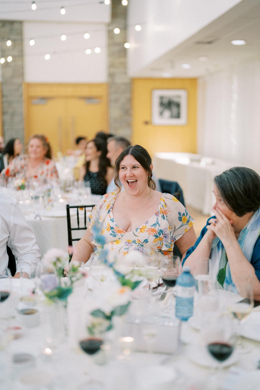A joyful moment captured at a formal event, with a woman in a floral dress laughing among guests seated at elegantly set tables.