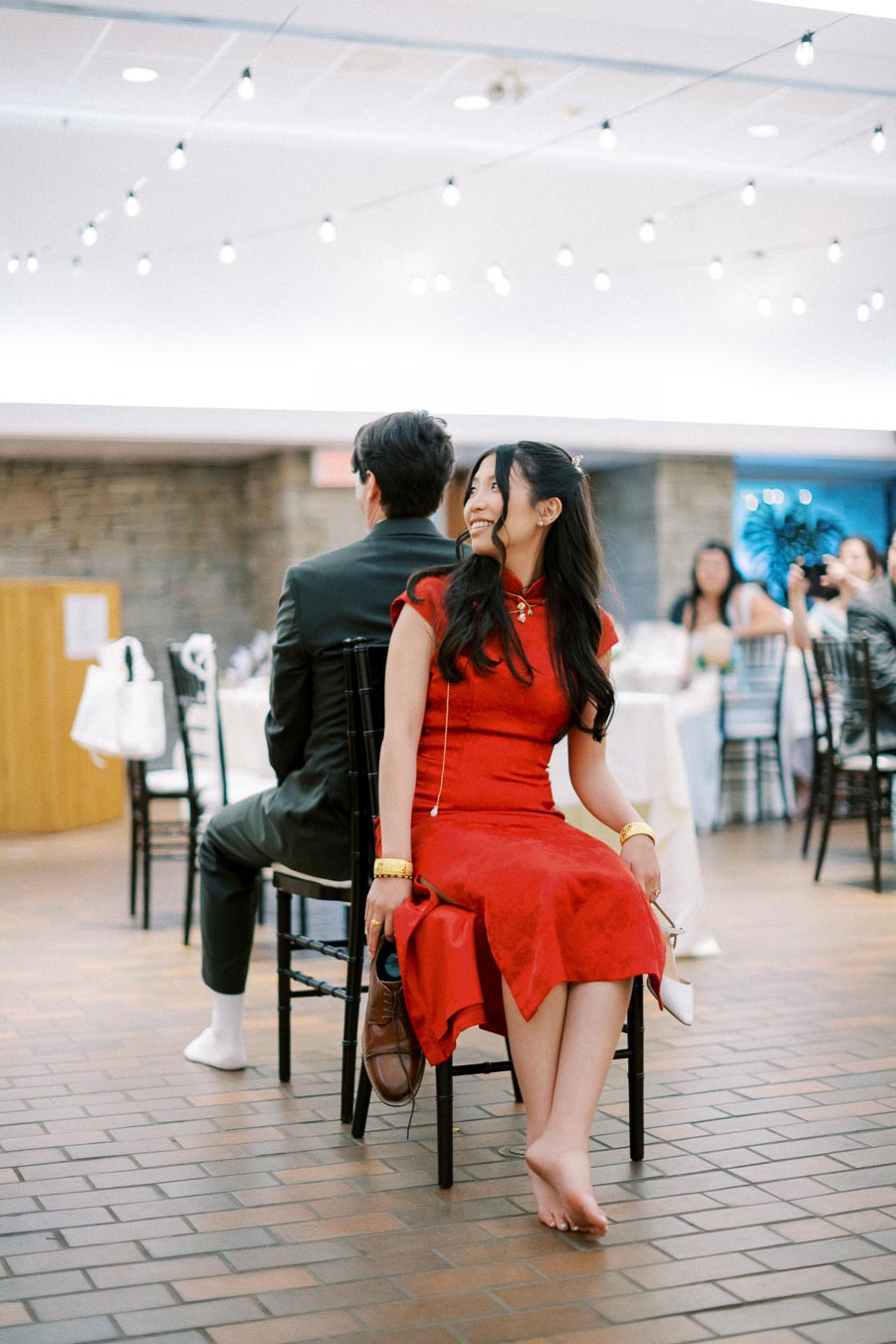 A woman in a red dress and a man in a suit participate in a fun activity at an indoor event, sitting back-to-back on chairs.