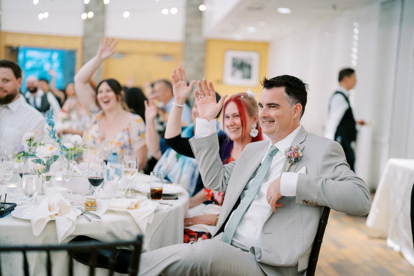 Guests at a wedding reception joyfully raise their hands during a celebration, seated at a table adorned with flowers and drinks.