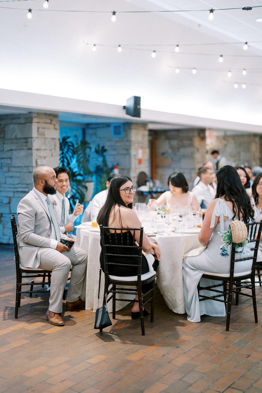 Guests enjoy a formal gathering, seated around a table adorned with elegant tableware, under ambient string lighting.