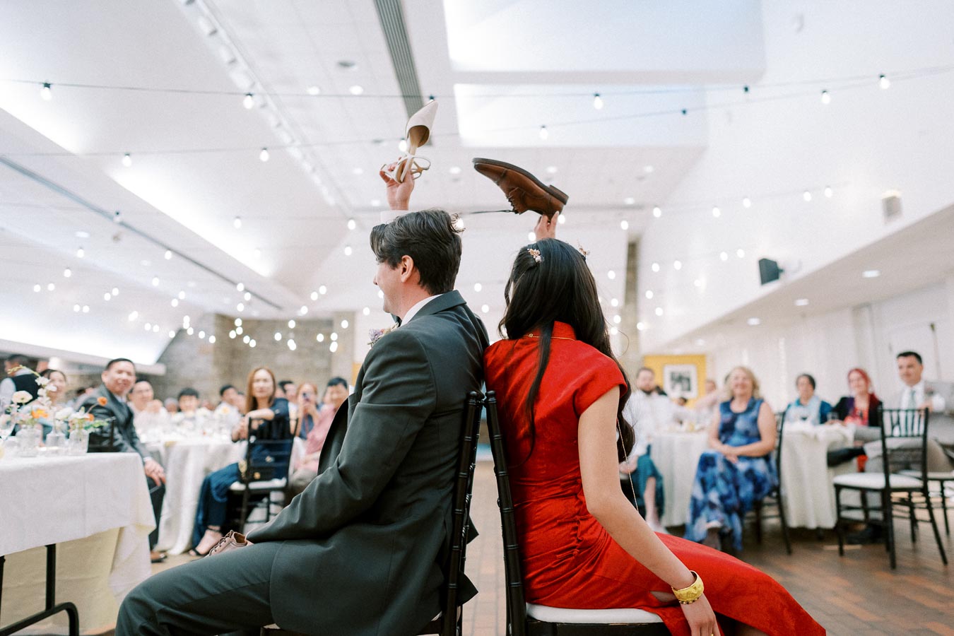 A couple participates in the shoe game during their wedding reception, sitting back-to-back and holding each other's shoes. Guests are seated around them, enjoying the event under string lights.
