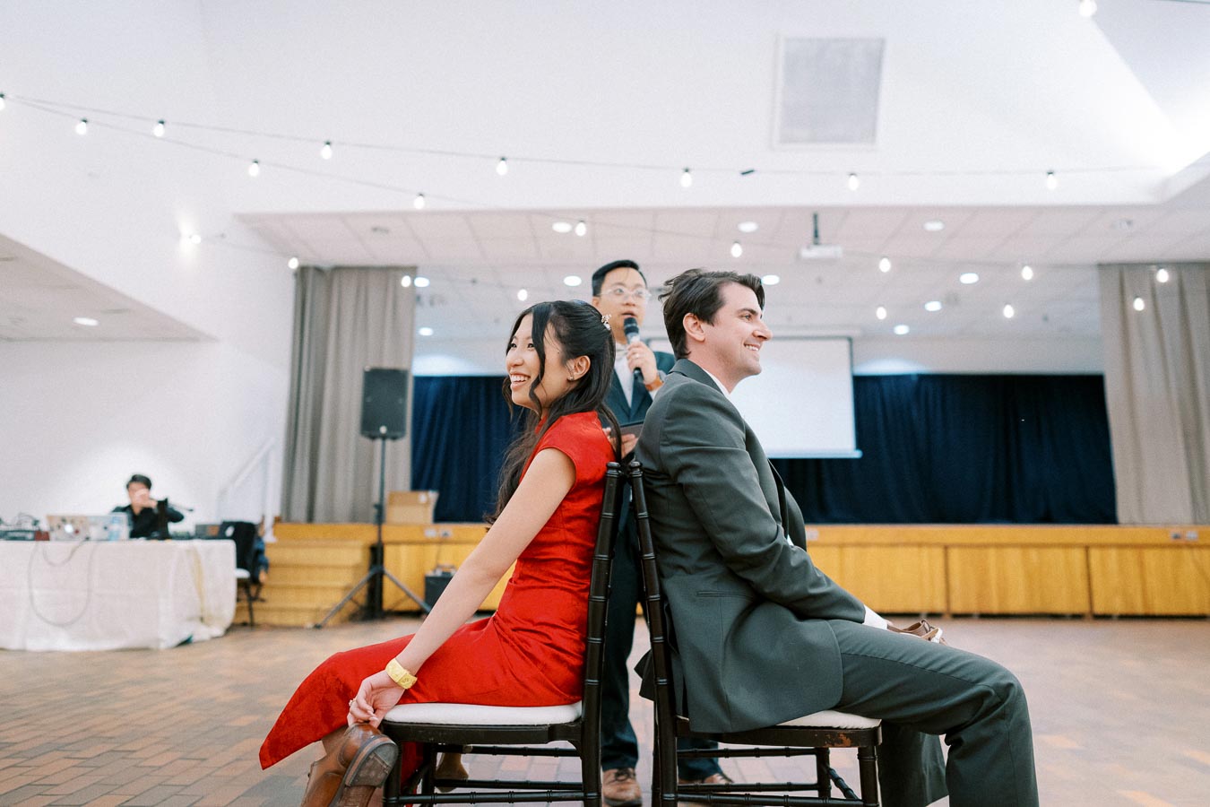 A couple participates in a fun activity, sitting back-to-back on chairs, while a speaker engages the audience in a decorated hall.