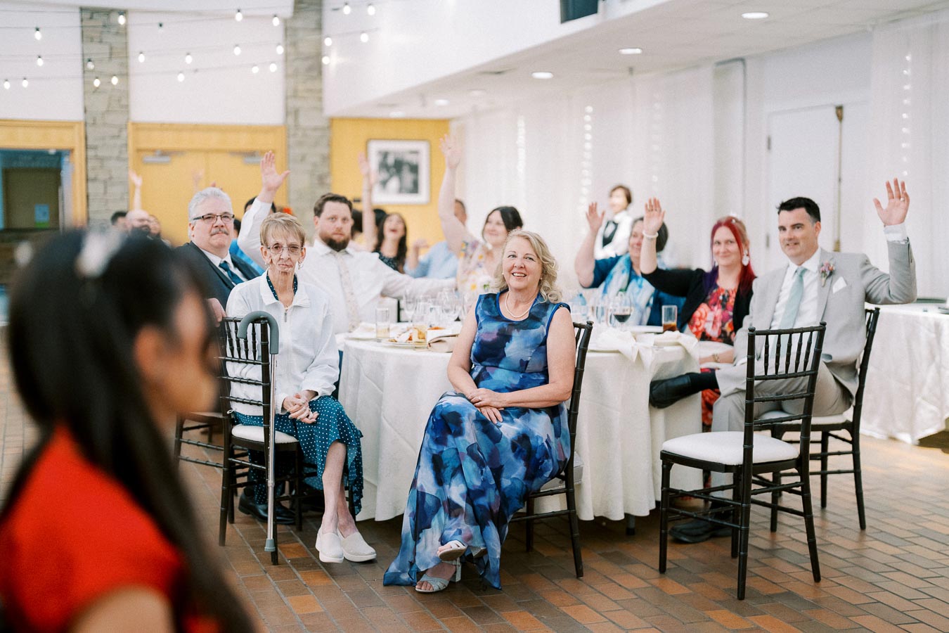 Guests at an indoor event, some raising their hands, seated around a table with white tablecloths and black chairs.