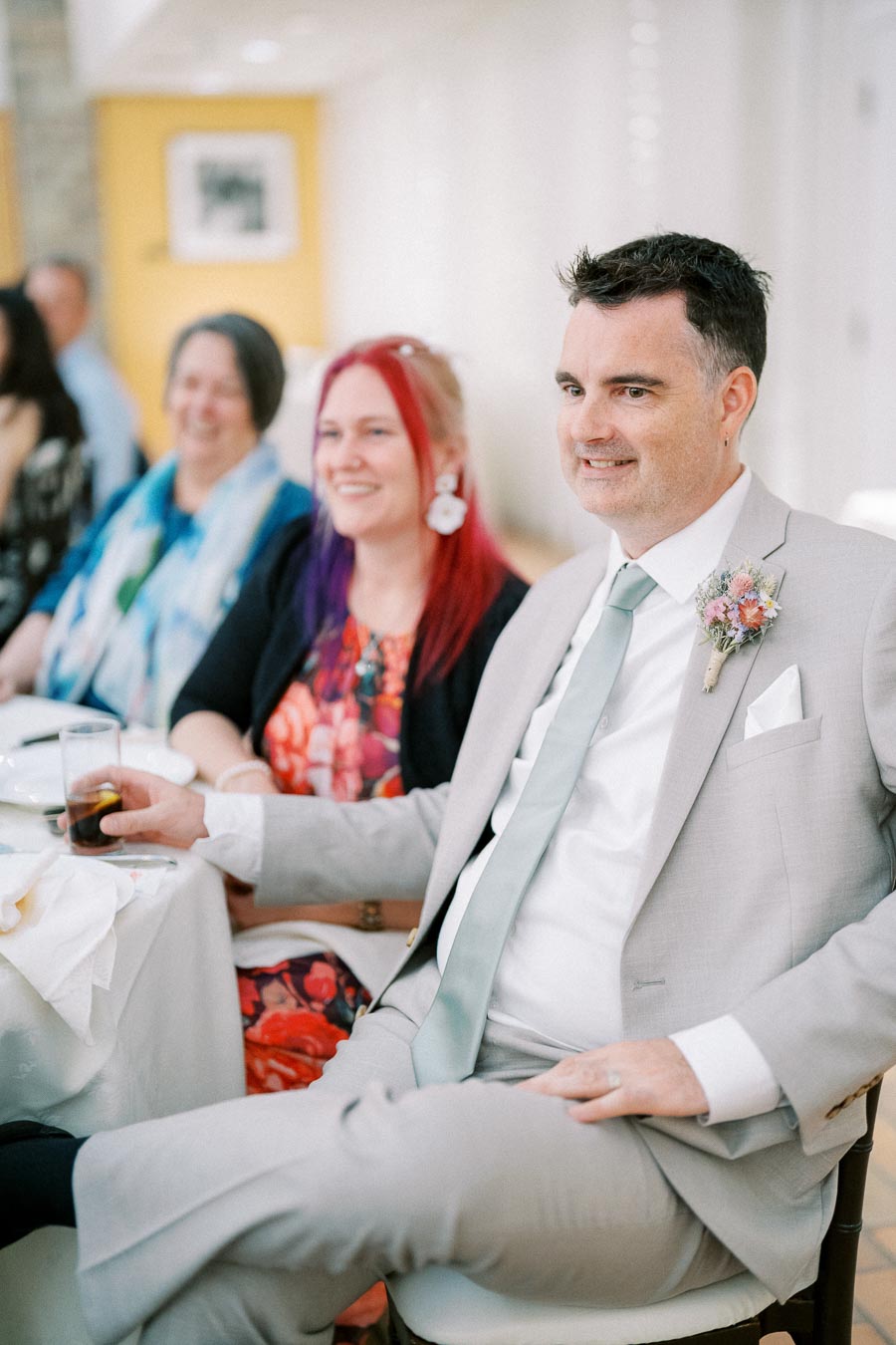 A man in a light gray suit with a floral boutonniere sits at a table with others, smiling at a formal event.