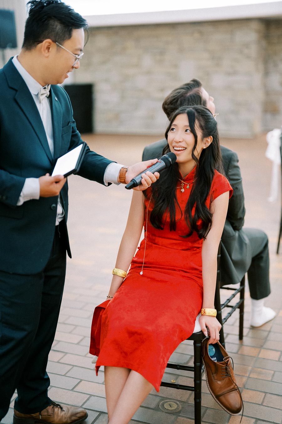 A woman in a red dress participates in an event, holding a shoe and speaking into a microphone held by a man in a suit.