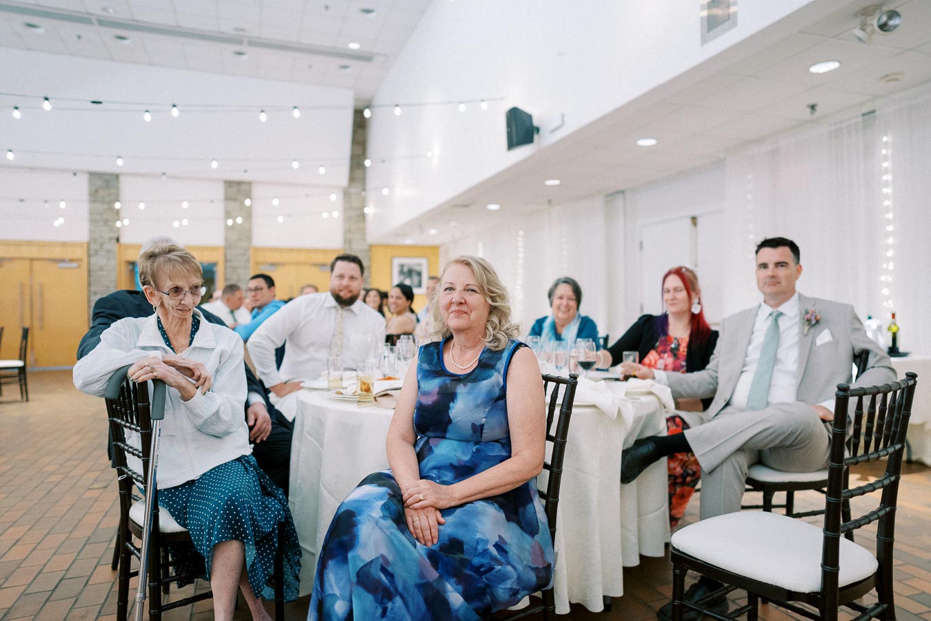 Guests attentively listening during a formal event, seated around a table adorned with glasses and plates.