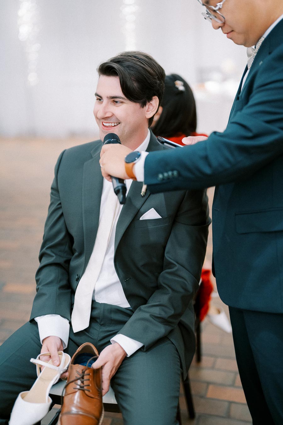 A man in a suit holds a microphone and two shoes, one brown and one white, while smiling during an event.