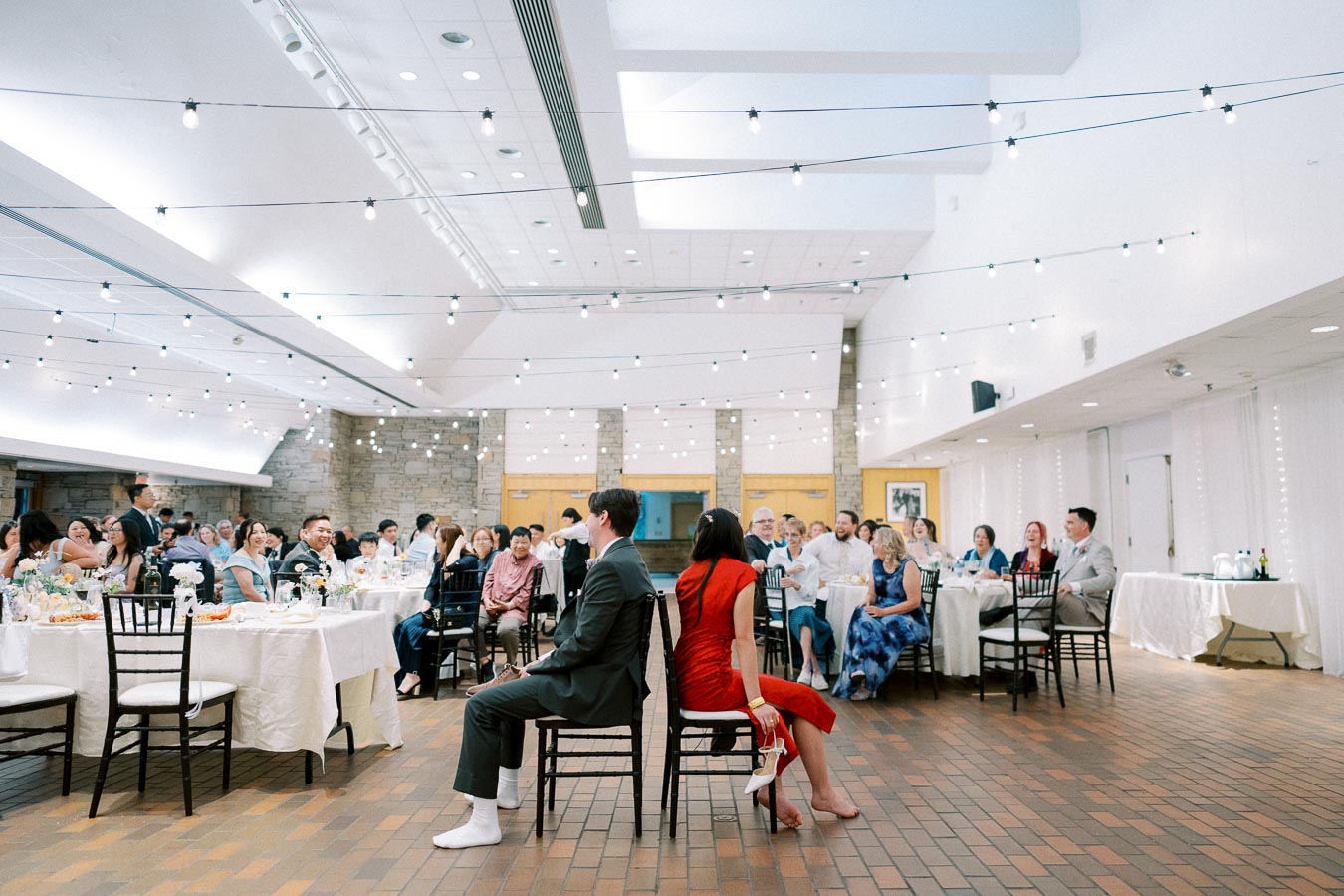 A couple sits back-to-back in chairs during a wedding reception game, surrounded by guests at tables under string lights in a spacious, decorated venue.