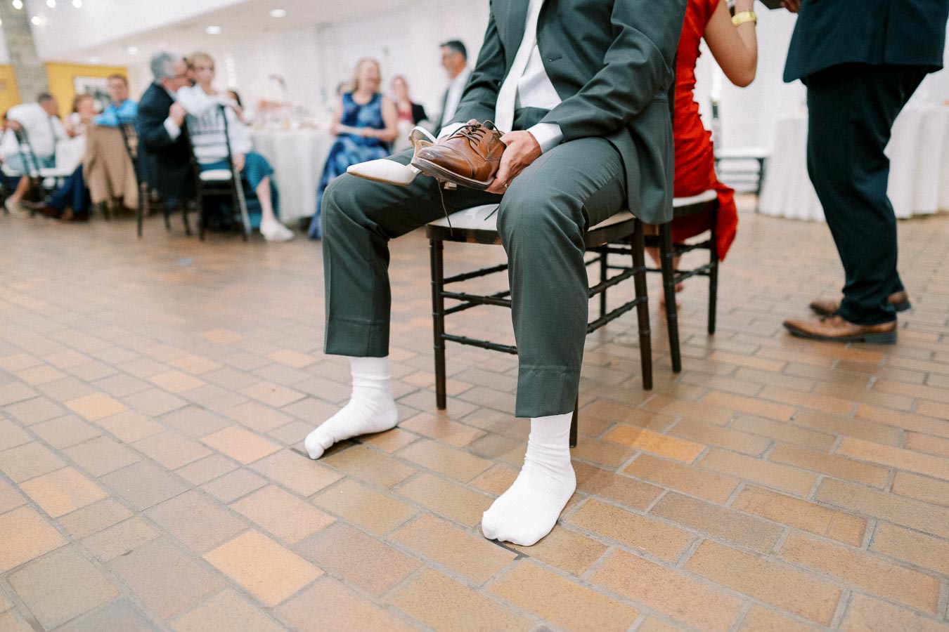 A man wearing a suit and white socks, sitting in a chair at a festive gathering, holding brown shoes during a wedding game with guests in the background, capturing a fun moment of the celebration.