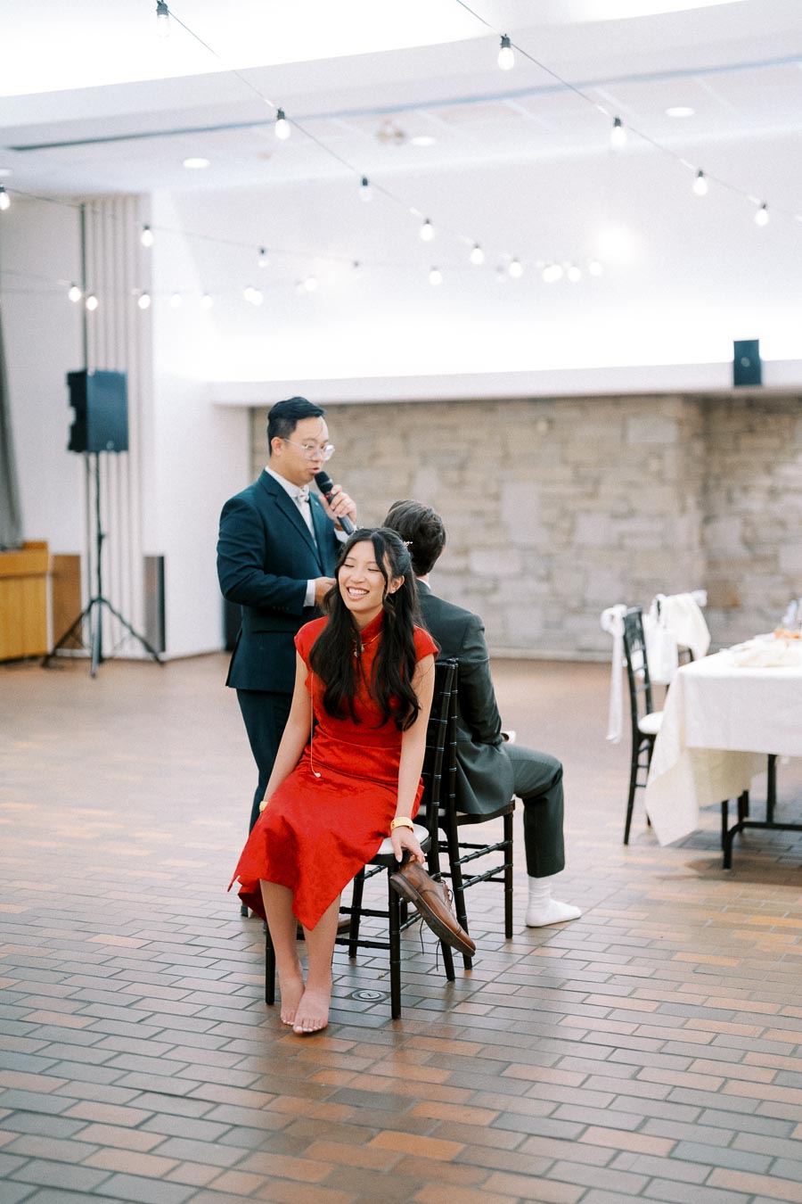 A joyful woman in a red dress smiles during a wedding game, seated back-to-back with a partner on chairs, while a man in a suit speaks into a microphone, under string lights in a spacious venue.