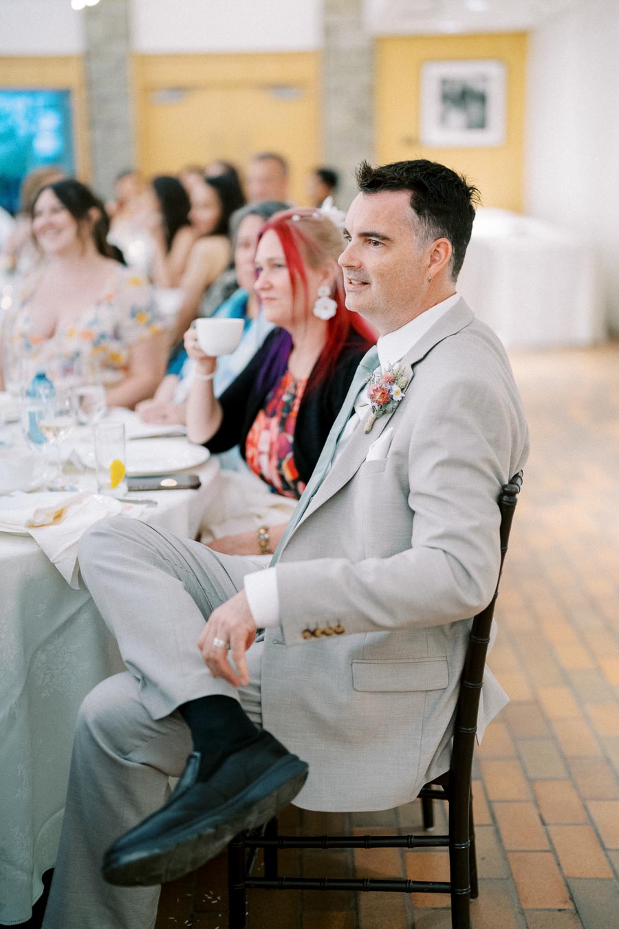 A man in a light gray suit sits at a wedding reception, attentively watching a speech. The table is set with glasses and plates, and other guests are visible in the background, dressed in formal attire. The atmosphere is festive and elegant.