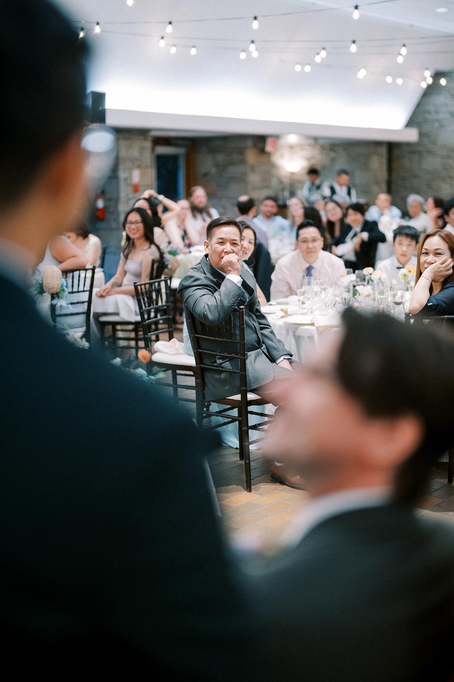 A thoughtful wedding guest in a suit listens attentively during a reception speech, surrounded by elegantly dressed attendees seated at decorated tables in a warmly lit venue.