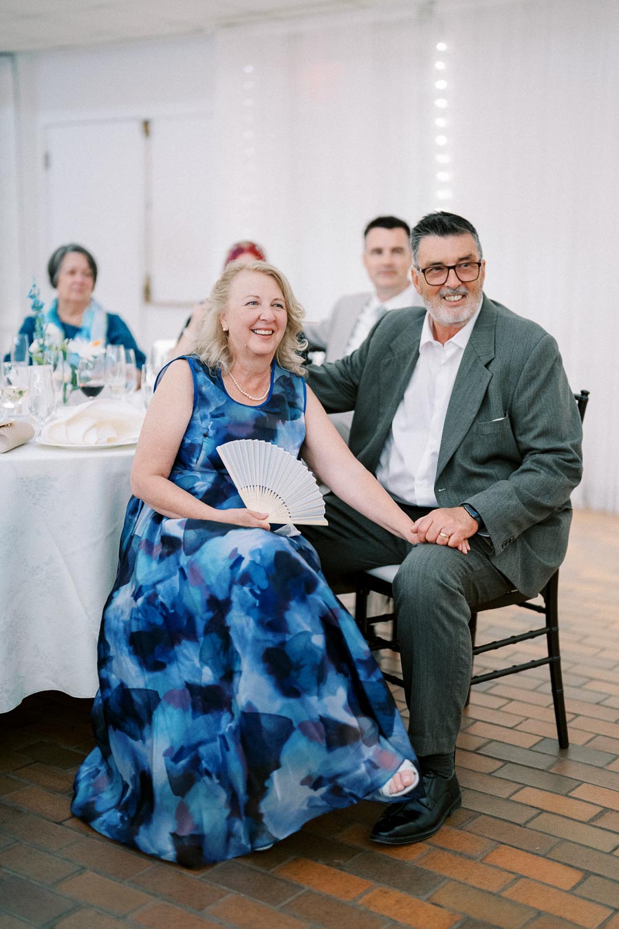 Elderly couple at an event, woman in blue floral dress holding a fan, smiling while seated next to a man in a grey suit, with people in the background.