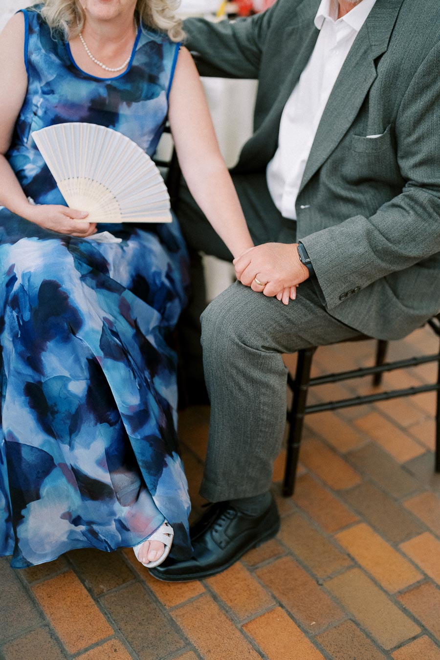 A couple sitting closely, holding hands; woman in a blue floral dress with a fan, and man in a gray suit on a brick patio.