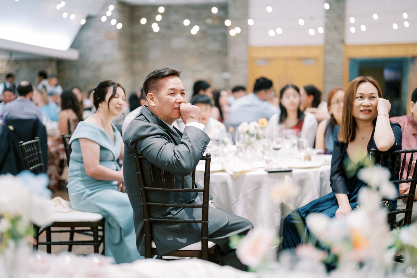 A joyful wedding reception with guests seated at elegantly decorated tables, adorned with floral centerpieces and illuminated by string lights.