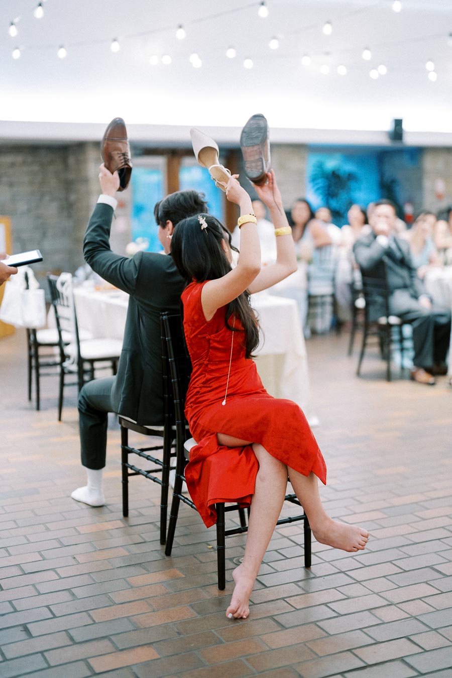 A bride in a red dress and a groom in a dark suit play the shoe game at a wedding reception, each holding up shoes while seated back-to-back on chairs in a warmly lit venue with guests watching.