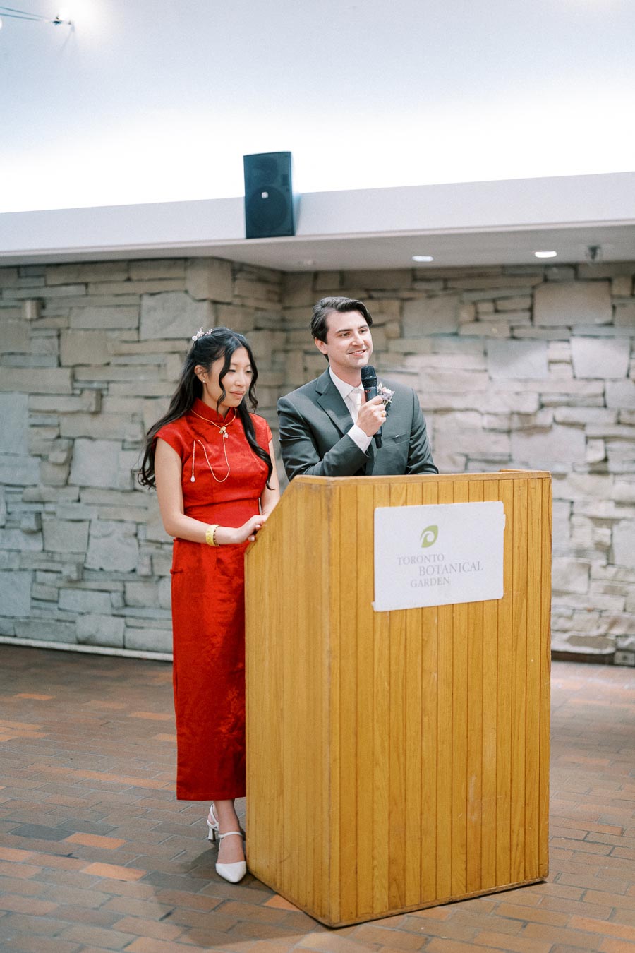 Two individuals stand together at a wooden podium with a sign reading Toronto Botanical Garden. The woman in a red dress and the man in a suit are speaking into a microphone, set against a stone wall backdrop, during an event or presentation.