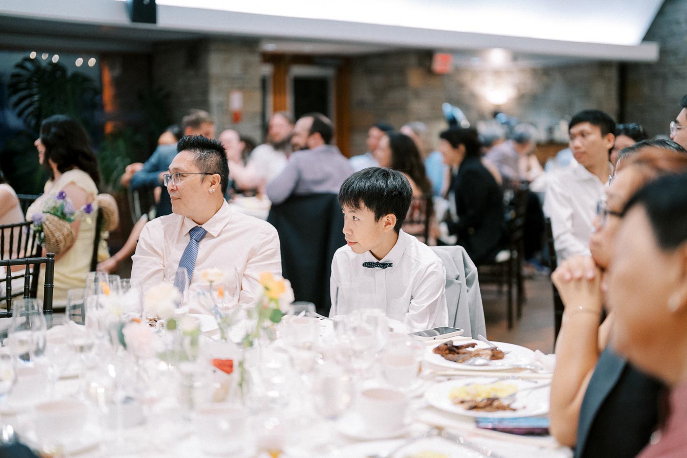 A diverse group of people seated at a decorated table during a formal event, engaged in conversation, with the focus on two individuals wearing shirts and ties, surrounded by elegantly arranged dinnerware and floral centerpieces.
