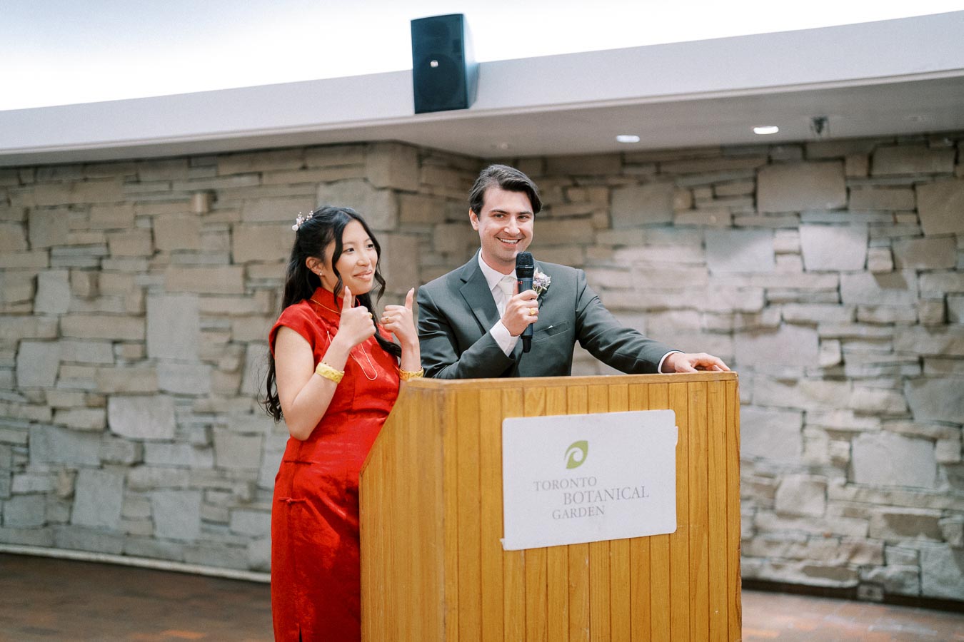 A couple speaking at a podium inside Toronto Botanical Garden, with the woman wearing a traditional red dress and giving a thumbs-up, while the man holds a microphone and smiles.