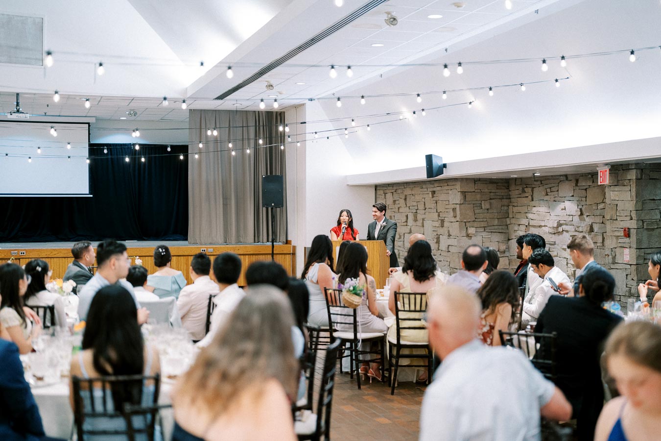 A lively indoor wedding reception with guests seated at tables, focused on two speakers at a podium, under elegant string lighting, against a modern stone wall backdrop.