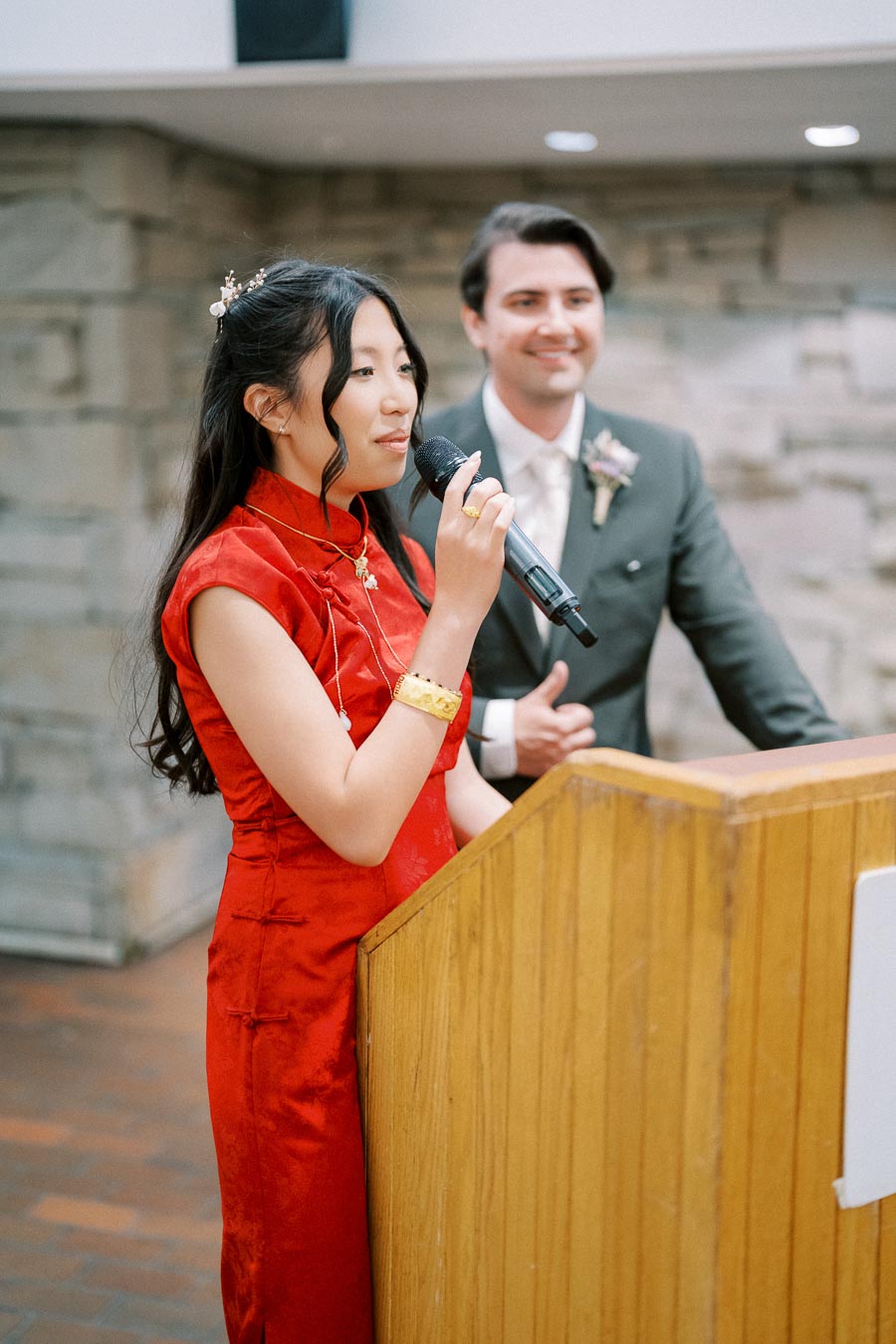 A woman in a red traditional dress speaks into a microphone at a podium, with a man in a suit standing beside her, indoors with a stone wall backdrop.