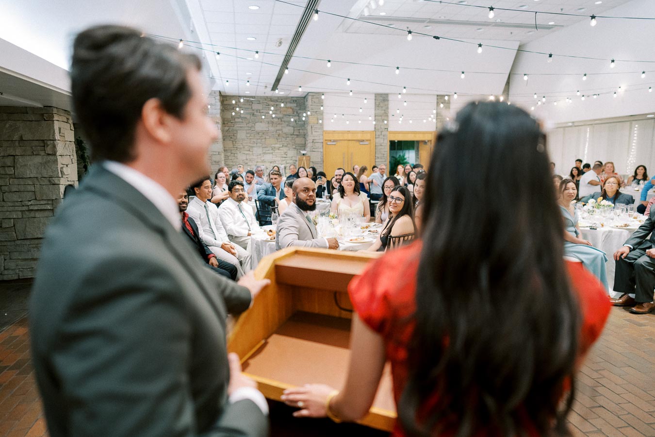 People gathered in a decorated event hall, listening to presenters at a podium during a festive occasion.