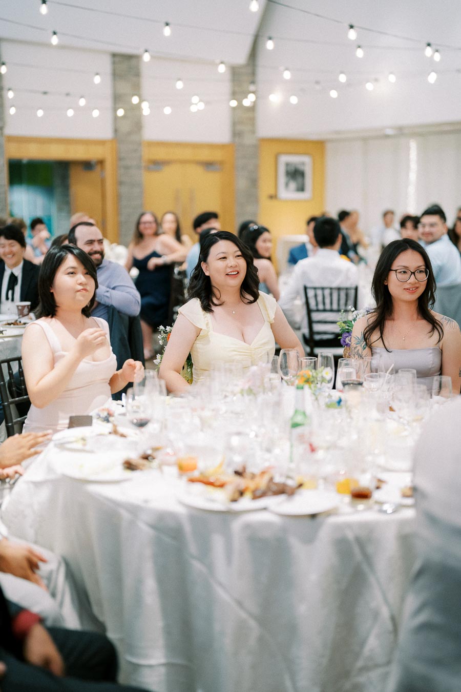 A group of guests enjoying a celebratory banquet in a decorated reception hall, with elegantly dressed individuals sitting around a table adorned with plates and glasses. The ambiance is lively, highlighted by hanging string lights and joyful expressions.