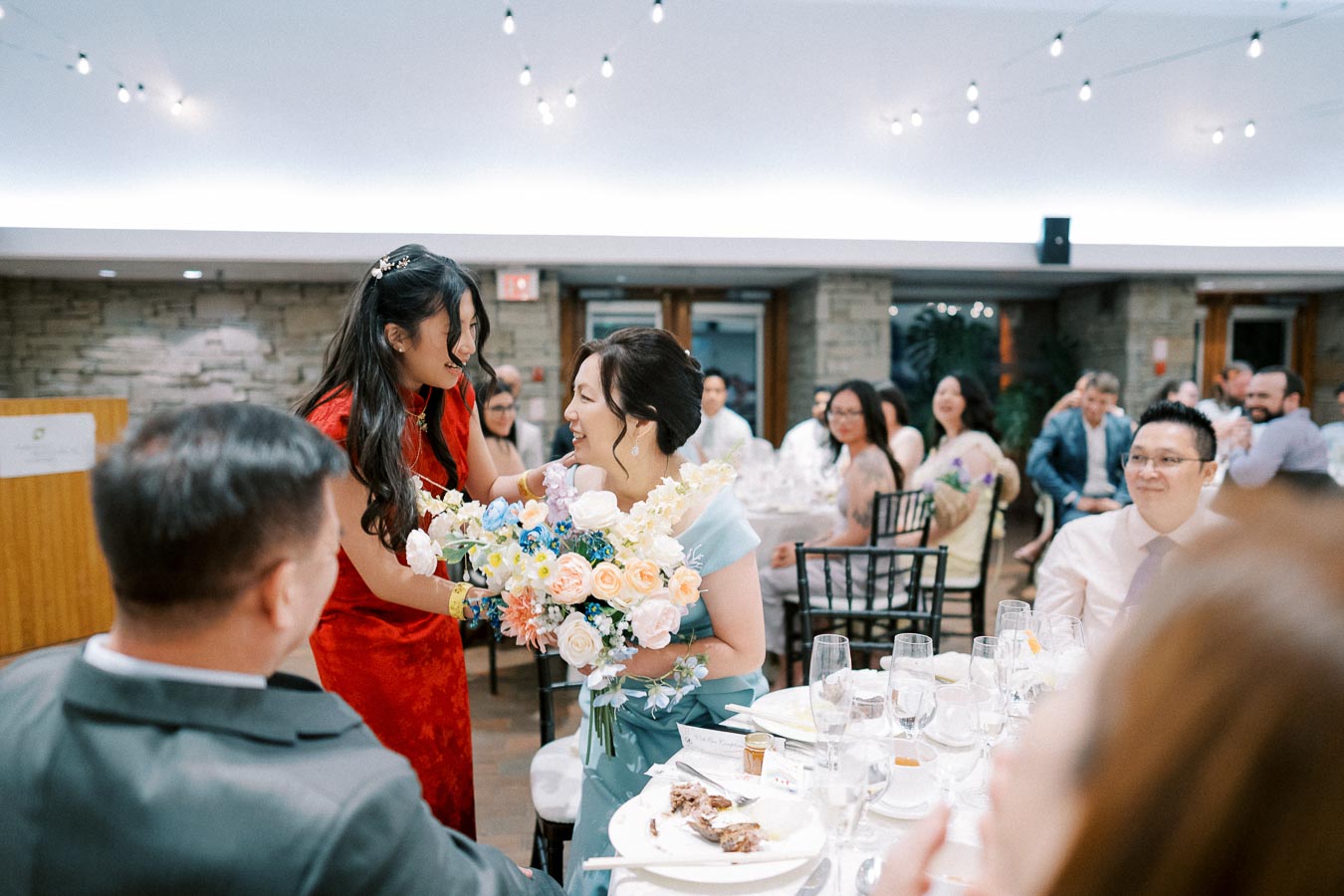 A joyful wedding reception with guests laughing and celebrating, featuring a woman in a red dress handing a colorful bouquet to a seated woman amidst elegantly set tables, captured under softly glowing string lights.