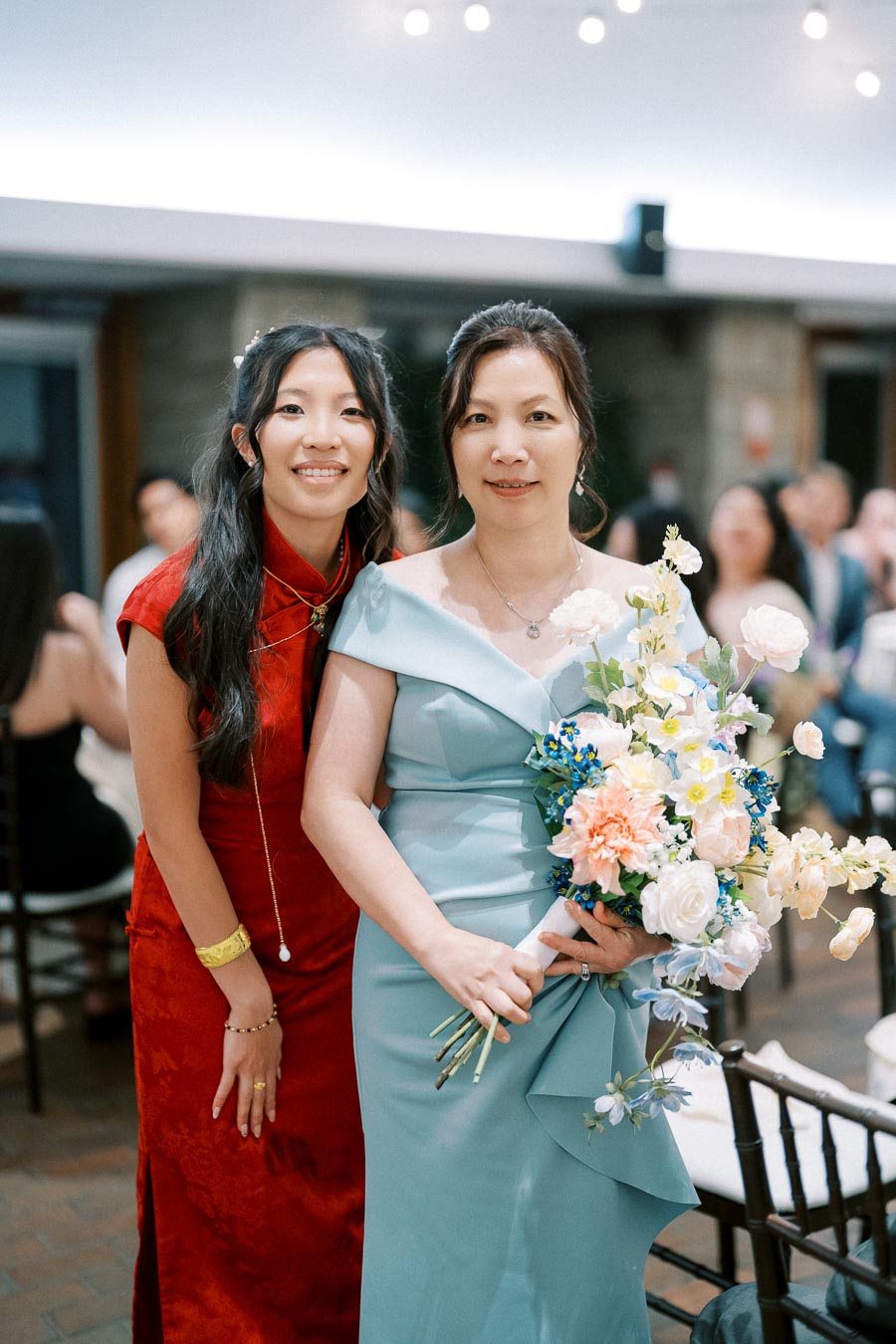 Two women smiling at a wedding reception, one in a red dress and the other holding a bouquet in a light blue gown, with party lights in the background.