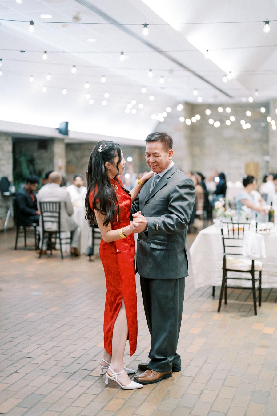 Father and daughter dancing at an elegant wedding reception, with the daughter in a red dress and the father in a suit. The venue features warm ambient lighting and guests seated at tables in the background.